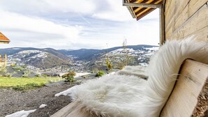 A wooden bench with a white furry cushion, overlooking a snowy landscape with buildings and mountains.