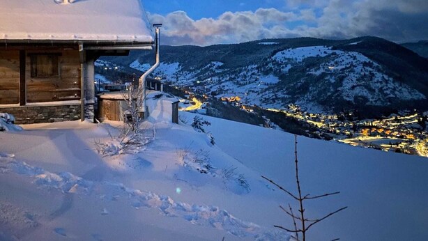 A snow-covered mountain landscape at night with a cabin and a lit town in the background.