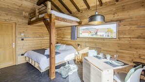 A wooden cabin with a bunk bed, a desk with a chair, and a window with a view of a building.