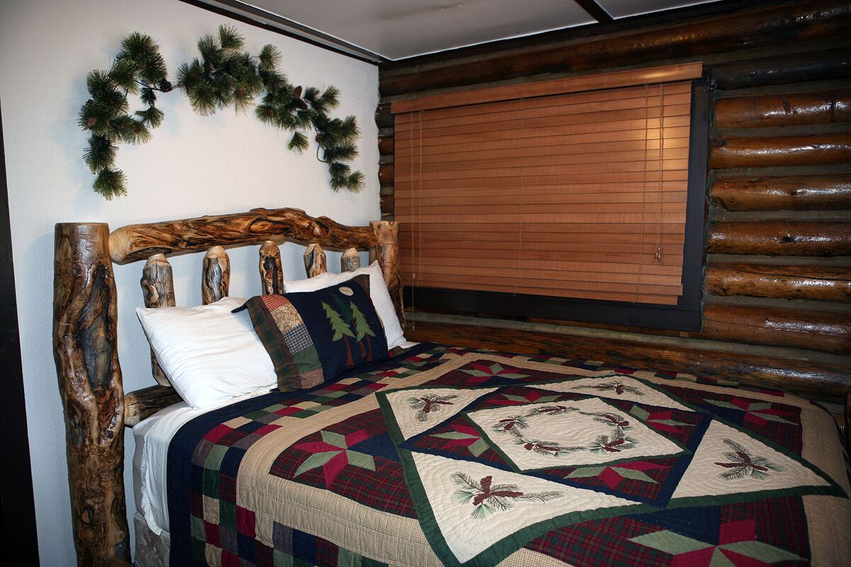 A bed with a wooden headboard, a quilt with geometric and floral patterns, and a window with blinds.