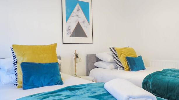 Une chambre d’hôtel avec deux lits, chacun agrémenté d’un oreiller bleu et jaune, d’une tête de lit blanche et d’une couverture turquoise. Un tableau encadré est accroché au mur et un radiateur se trouve sous les lits.
