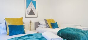A hotel room with two beds, each with a blue and yellow pillow, a white headboard, and a teal blanket. There is a framed picture on the wall and a radiator below the beds. - Tudors eSuites Birmingham House with Private Garden Free Parking (Birmingham)