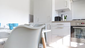 Une cuisine moderne avec des armoires blanches, un four encastré et un coin repas comprenant une chaise blanche et des tasses bleues.