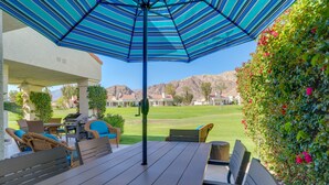 An outdoor patio with a striped blue umbrella, wicker furniture, and a view of a golf course and mountains. - PGA West Mtn Course: Luxury Golf Retreat! (La Quinta)