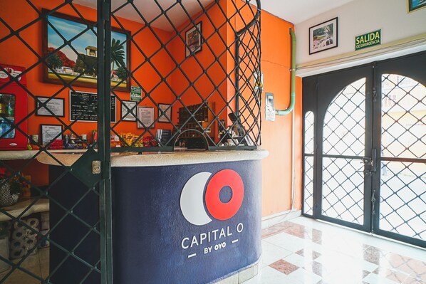 A reception area with a blue counter labeled "Capital O by OYO," a chain-link fence, and a door with a sign reading "Salida de Emergencia."