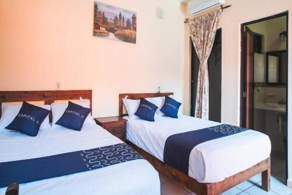 A hotel room with two beds, a painting on the wall, an air conditioning unit, and a bathroom visible through an open door.