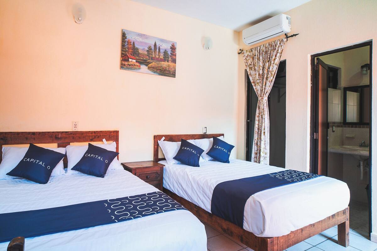 A hotel room with two beds, a painting on the wall, an air conditioning unit, and a bathroom visible through an open door.