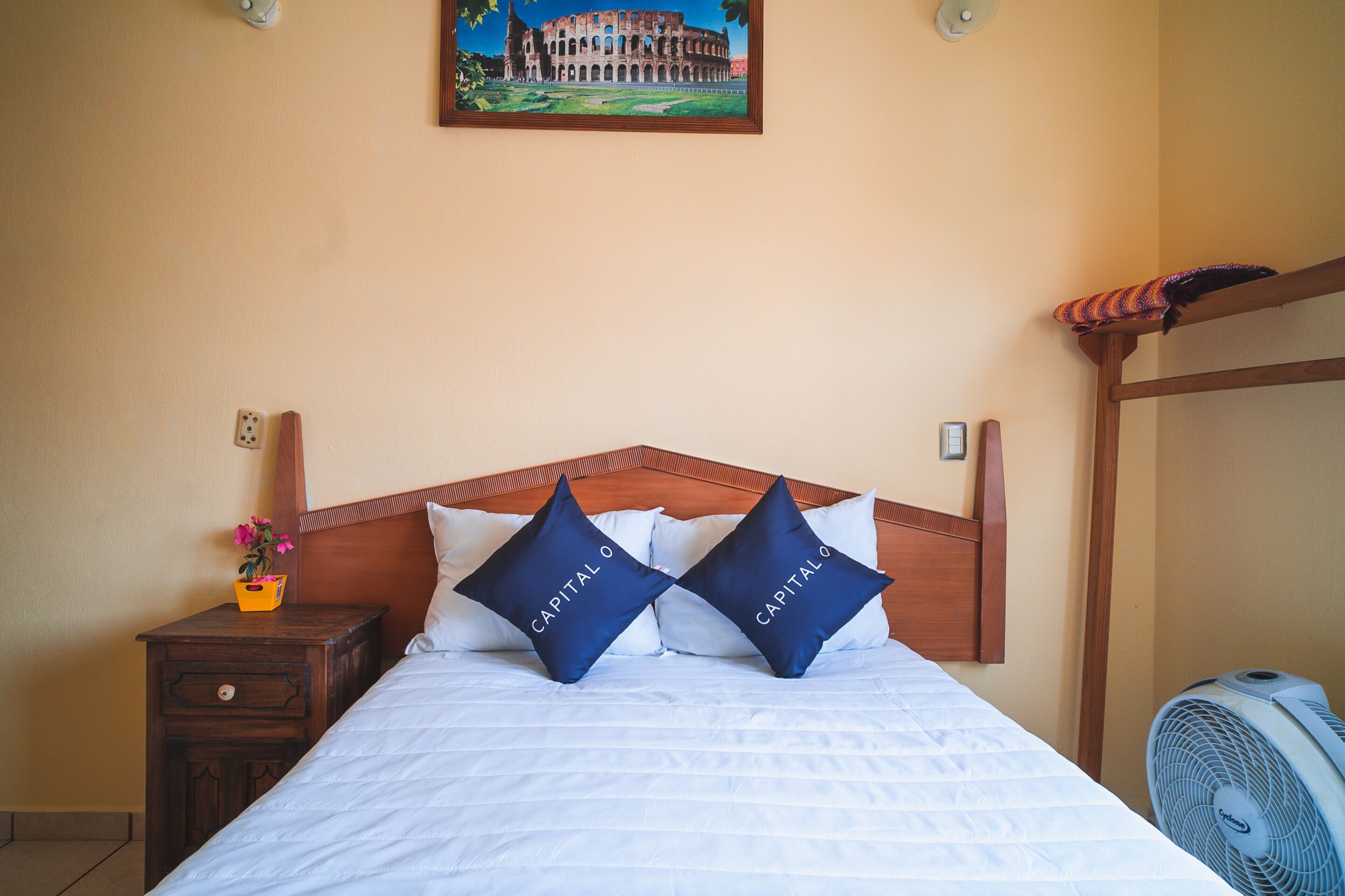 A neatly made bed with two pillows labeled "Capital O" in a hotel room with a wooden headboard, a nightstand with a vase of flowers, and a wall-mounted picture of a historical building.