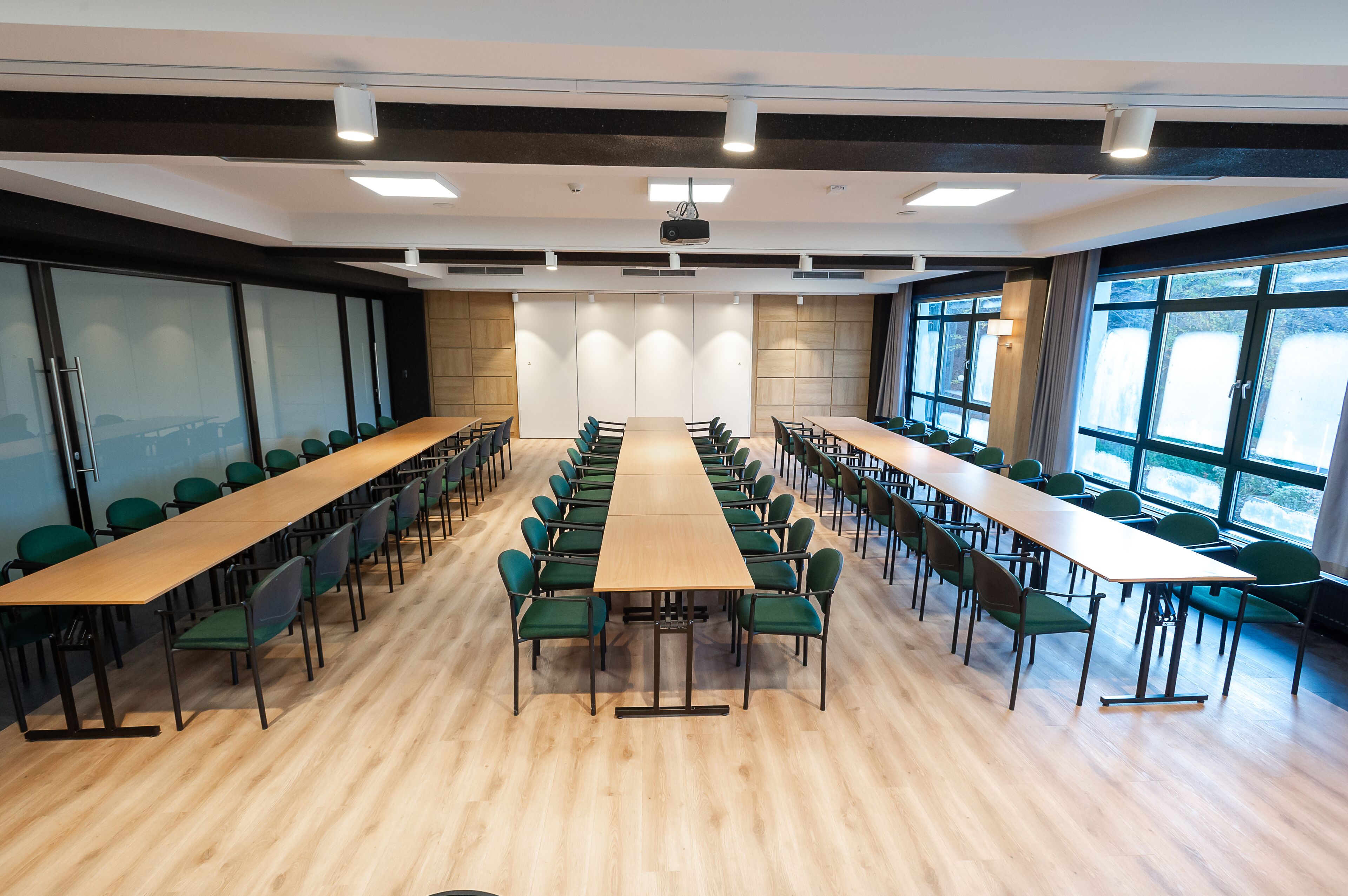 A conference room with long tables and chairs, large windows, and a projector on the ceiling.