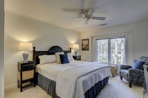 A bedroom with a bed, two bedside tables, a chair, a ceiling fan, and a large window. - 7653 Huntington - Beautiful Lagoon Views/Screened in Porch (Hilton Head Island)