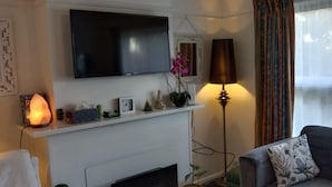Een woonkamer met een flatscreen televisie aan de muur, een open haard met schoorsteenmantel, een bank met decoratief kussen, en een vloerlamp.