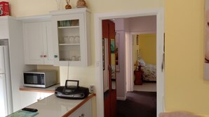 Een compacte keuken met een magnetron, een kleine koelkast en een kast met glazen. Een doorgang leidt naar een slaapkamer met een bed en een nachtkastje.