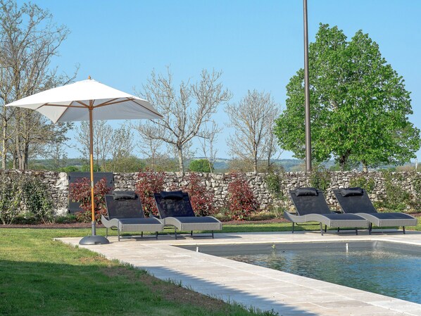 A pool area with lounge chairs and a large umbrella, surrounded by stone walls and greenery.