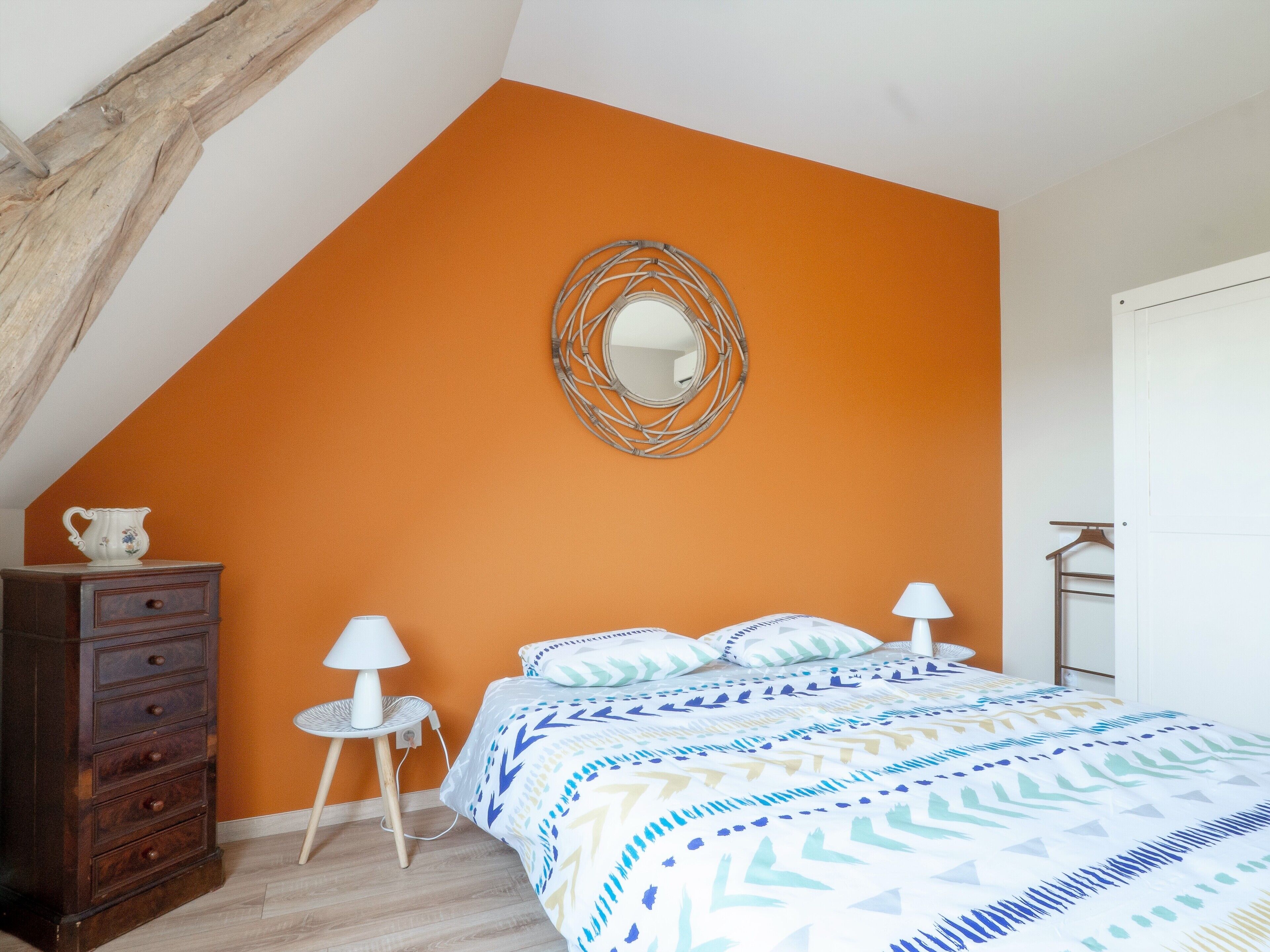 Une chambre avec un mur orange, un lit avec une literie à motifs, une commode, une petite table avec une lampe et un miroir sur le mur.