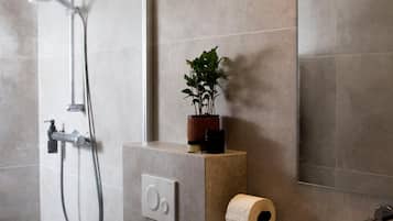 Un baño con ducha, un dispensador de papel higiénico y una planta en maceta sobre una estantería.