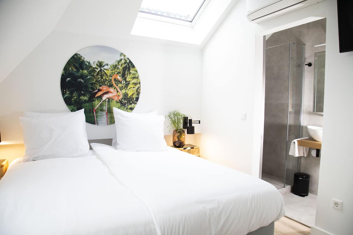 Una habitación de hotel moderna con tragaluz, una cama con ropa blanca, un adorno circular en la pared, zona de ducha y una planta pequeña en la mesita de noche.
