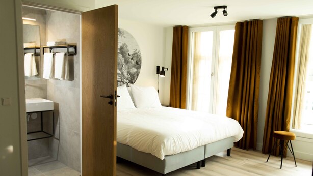 A bedroom with a bed, a bathroom with a sink and towel rack, and a large window with curtains. - Hotel Haverkist ('s-Hertogenbosch)