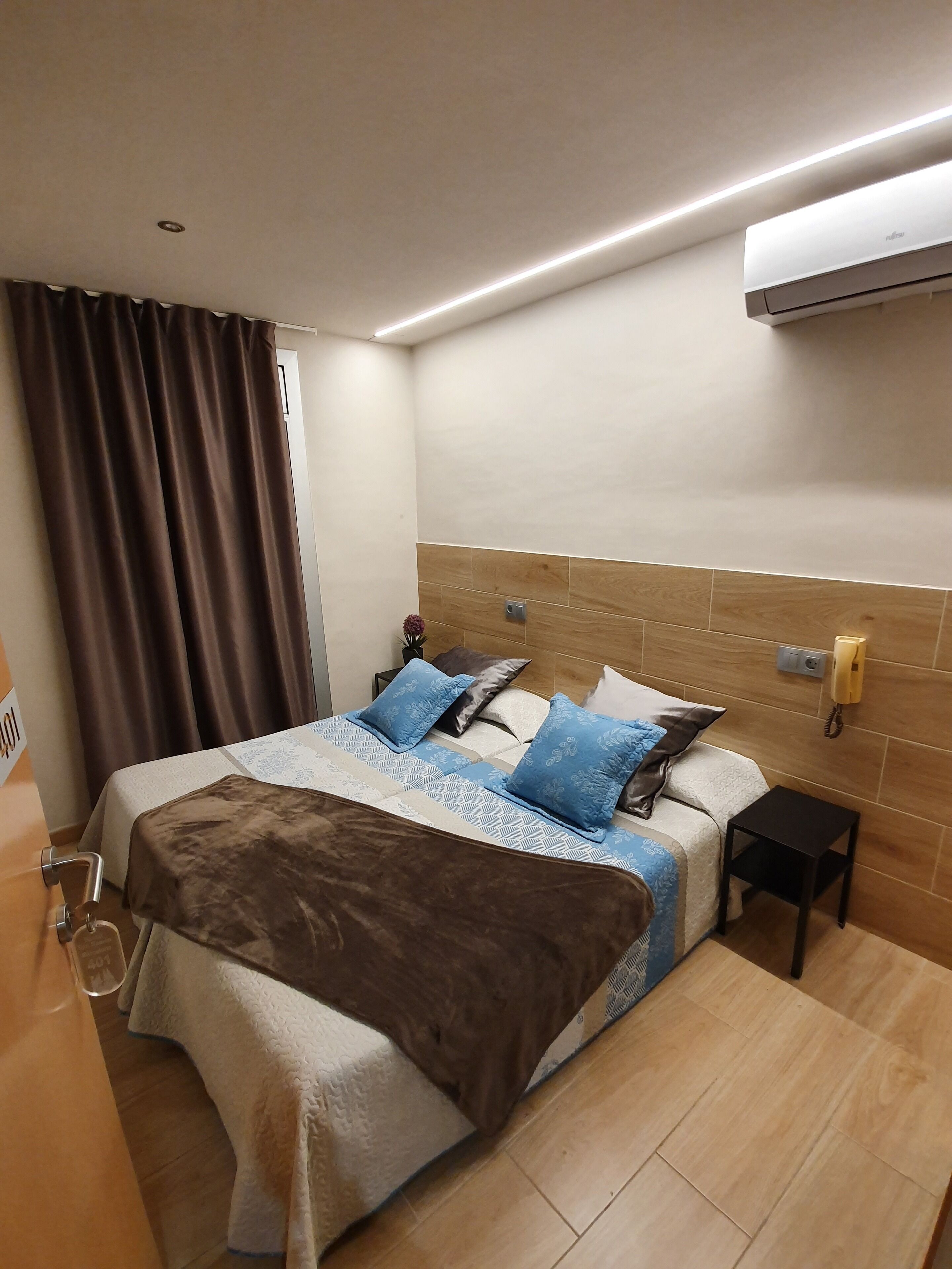 A hotel room with a bed, a bedside table, a nightstand, an air conditioning unit, and a curtain.