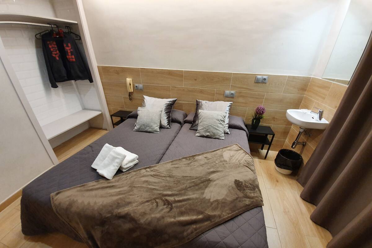 A hotel room with a bed, pillows, a bedside table with a lamp, a small sink, and a towel on the floor.