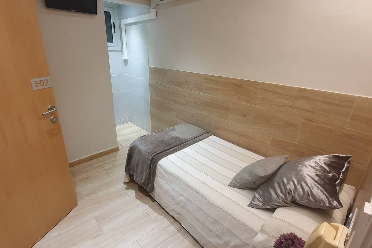 A hotel room with a bed, a bedside table with a telephone, and an air conditioning unit mounted on the wall.