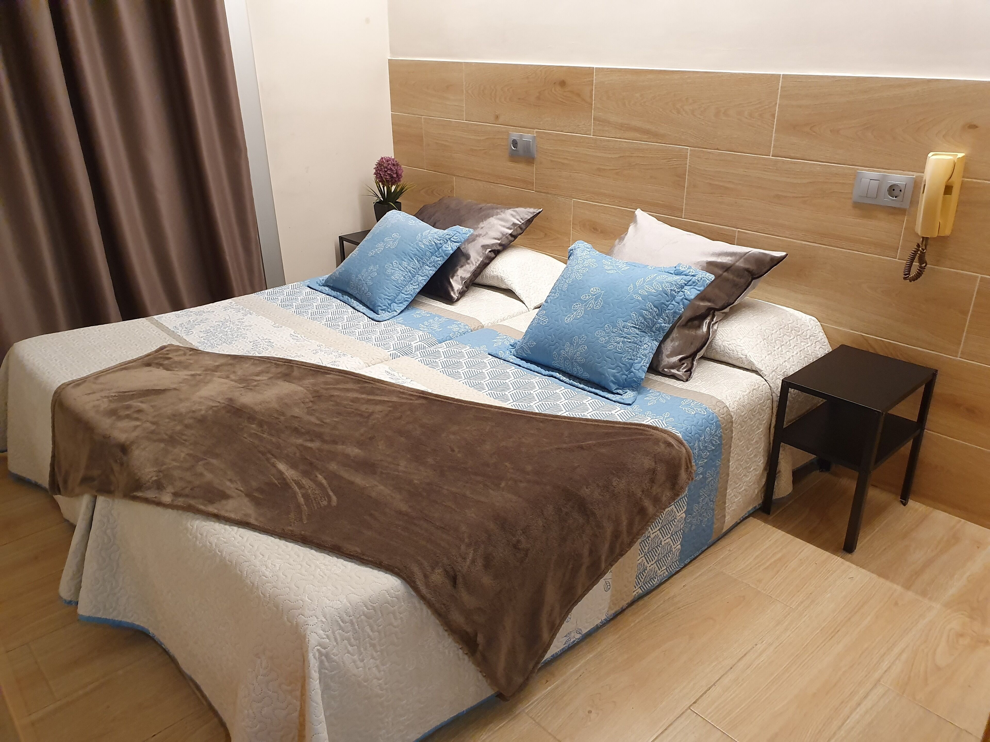 A neatly made bed with a brown blanket and patterned bedding, a bedside table with a lamp, and a wall-mounted phone.