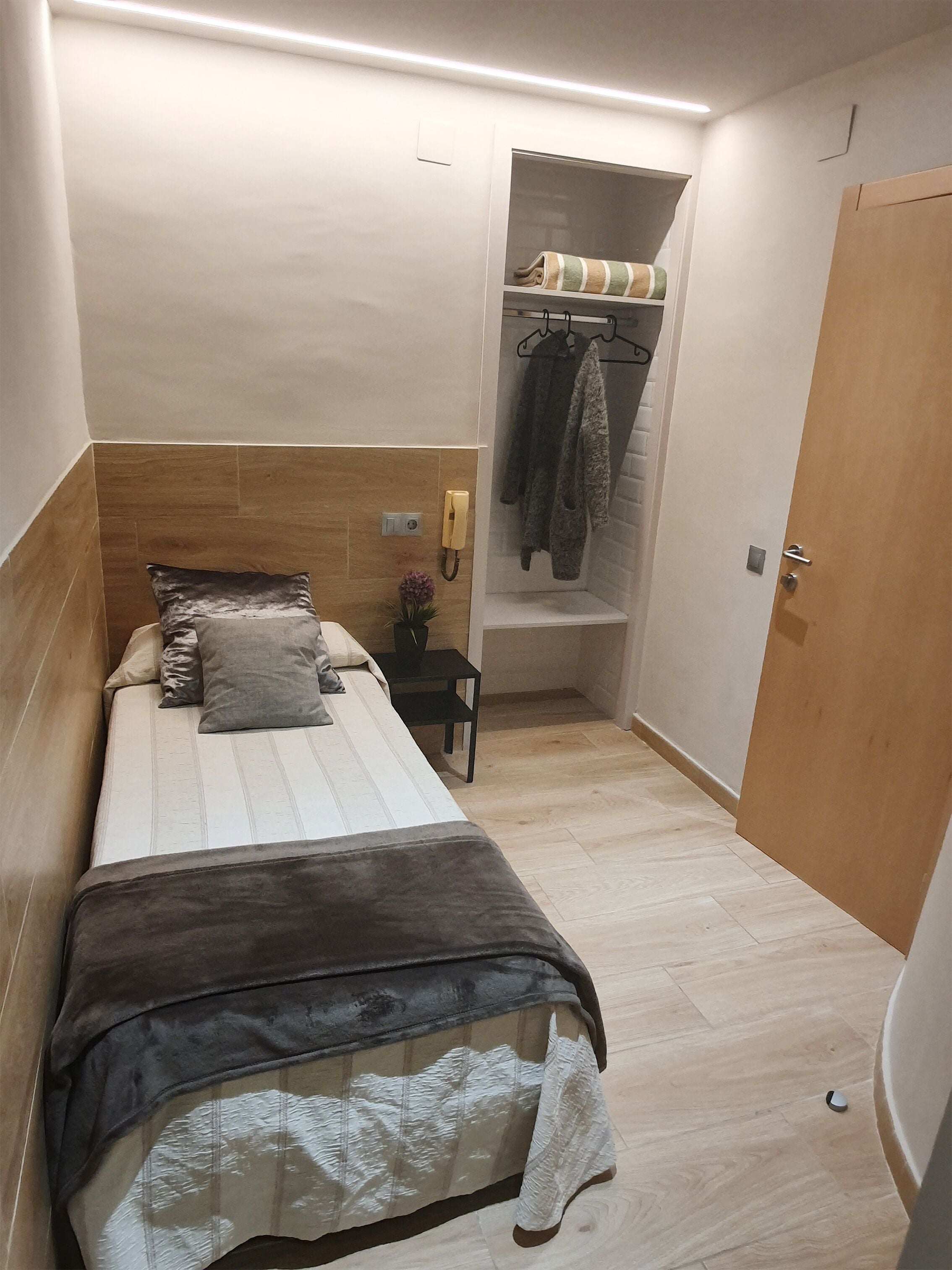A small, neatly arranged bedroom with a bed, a bedside table, a chair, a shelf with towels, and a door.