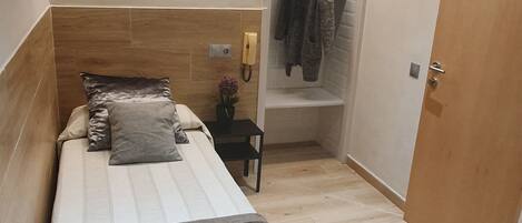 Un dormitorio pequeño y ordenado con cama, mesita de noche, silla, estante con toallas y una puerta.