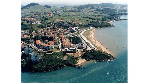 An aerial view of a coastal town with a beach, a fortified structure, and surrounding greenery.