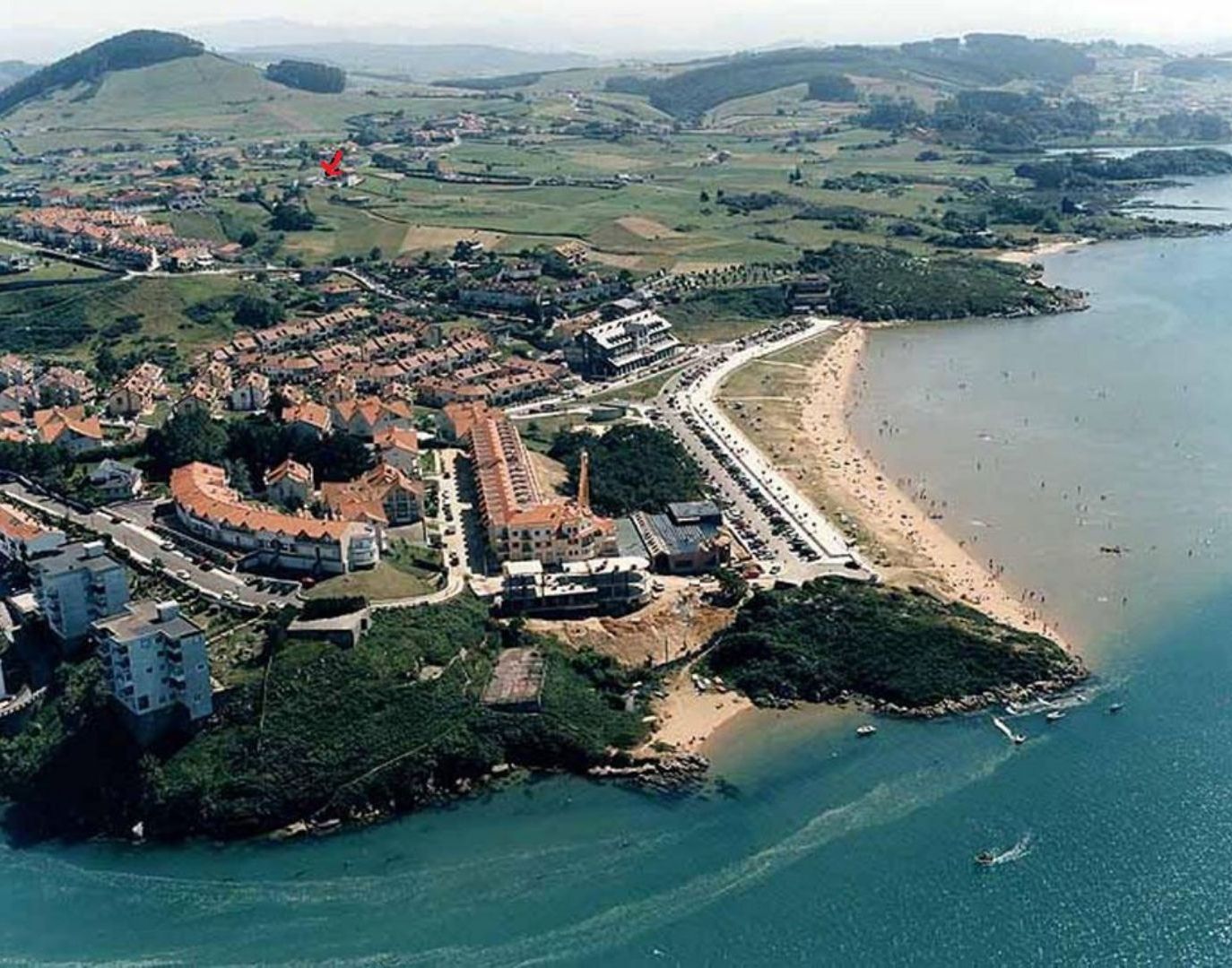 An aerial view of a coastal town with a beach, a fortified structure, and surrounding greenery.