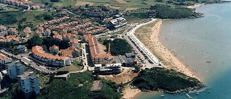 Vista aérea de uma cidade costeira com praia, estrutura fortificada e vegetação ao redor.
