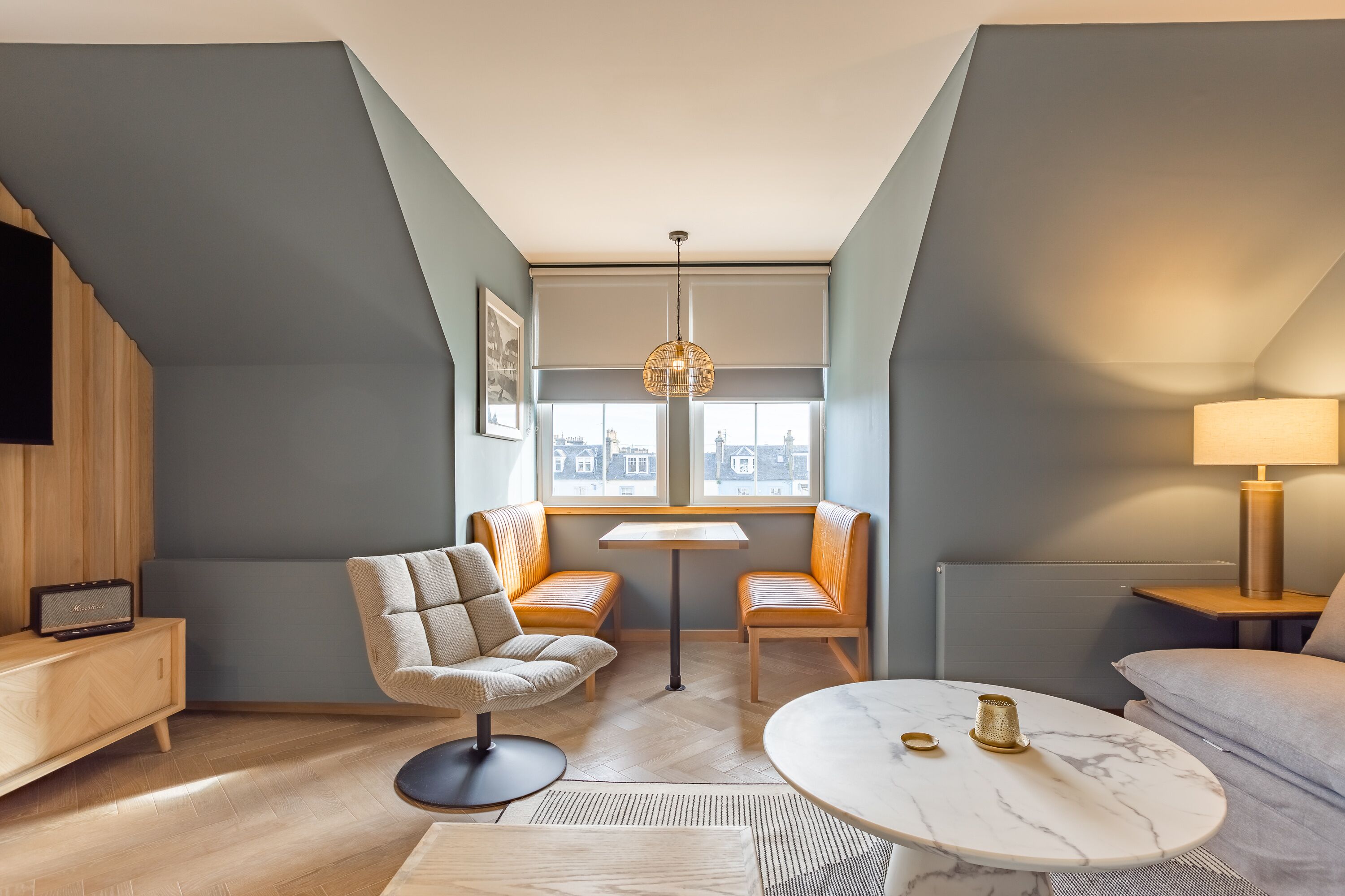 A modern hotel room with a loft design, featuring a sofa, a round table, a chair, and a television on a wooden cabinet.