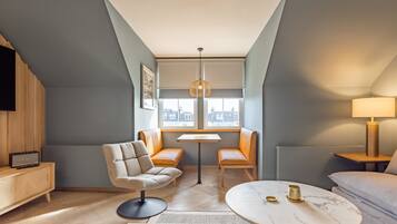 A modern hotel room with a loft design, featuring a sofa, a round table, a chair, and a television on a wooden cabinet.