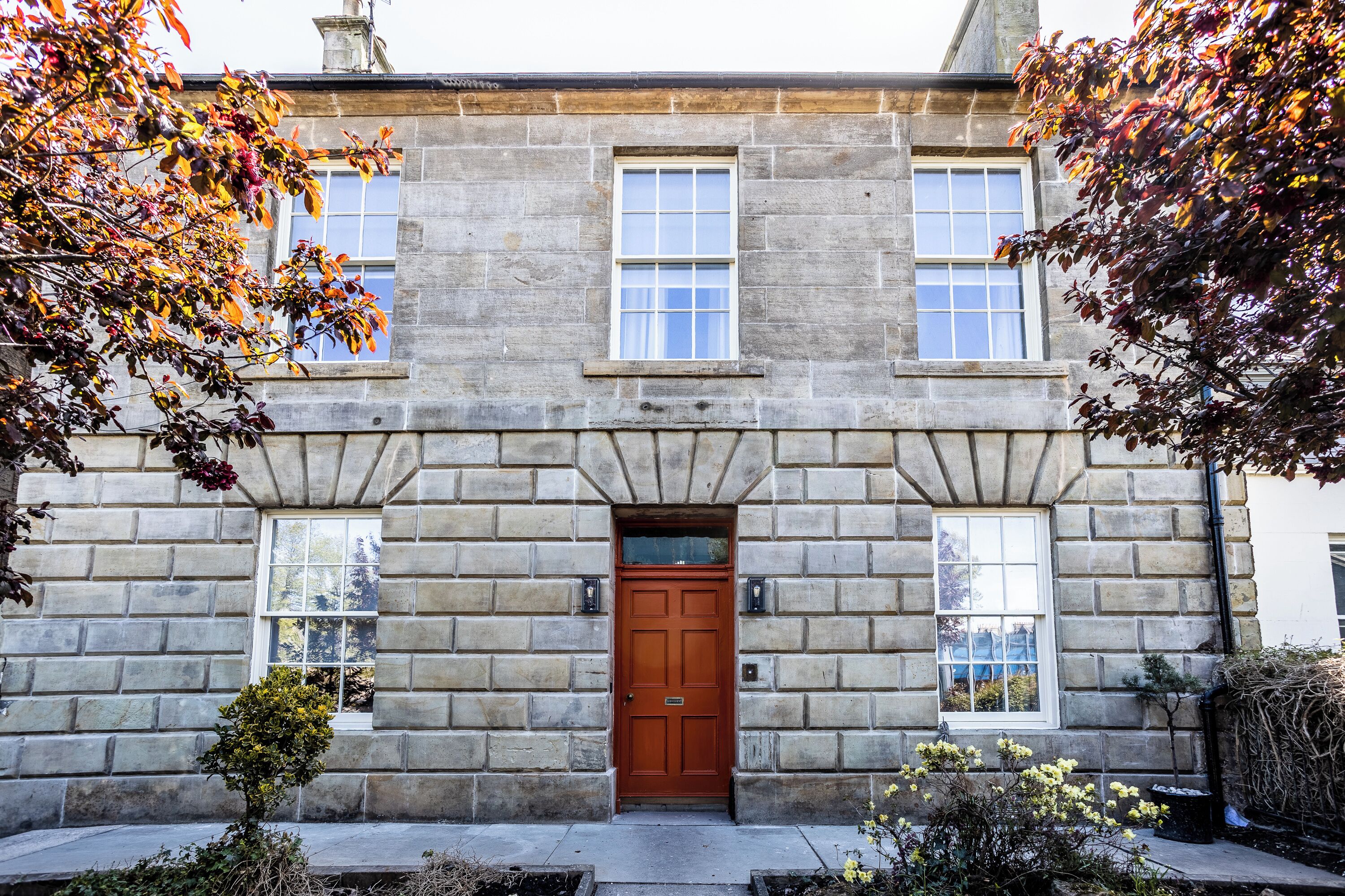 A stone building with a central entrance door, two windows on either side, and a small garden area with plants and a tree.