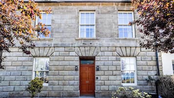 A stone building with a central entrance door, two windows on either side, and a small garden area with plants and a tree.