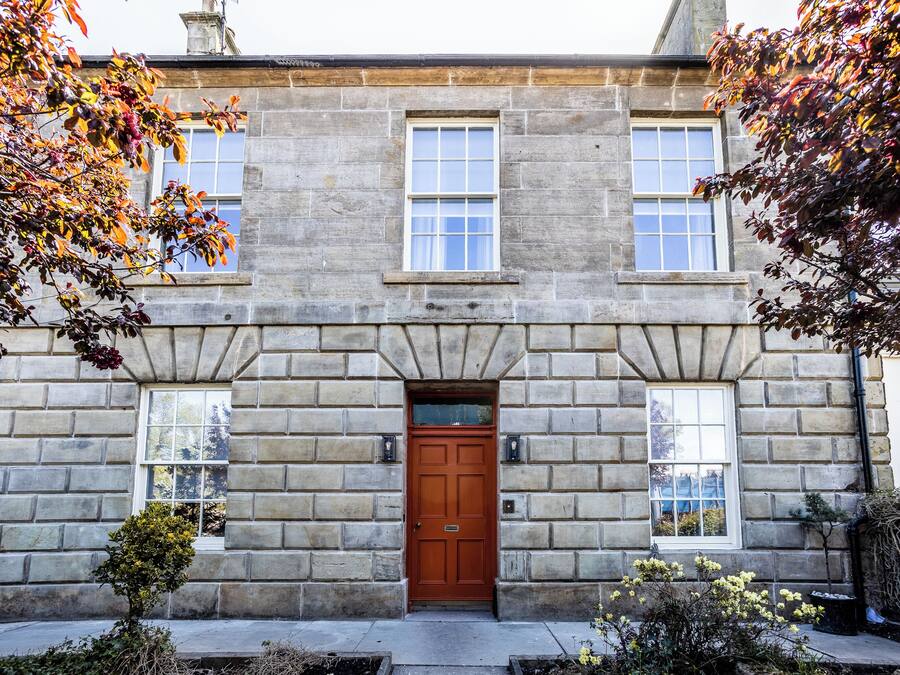A stone building with a central entrance door, two windows on either side, and a small garden area with plants and a tree.