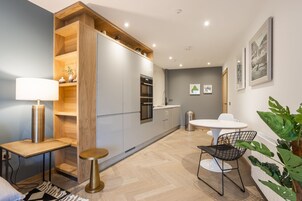 Una cocina moderna con armarios blancos, un horno empotrado y un estante de madera. Hay una mesa de comedor blanca con una silla negra, un taburete redondo pequeño y una lámpara de pie.