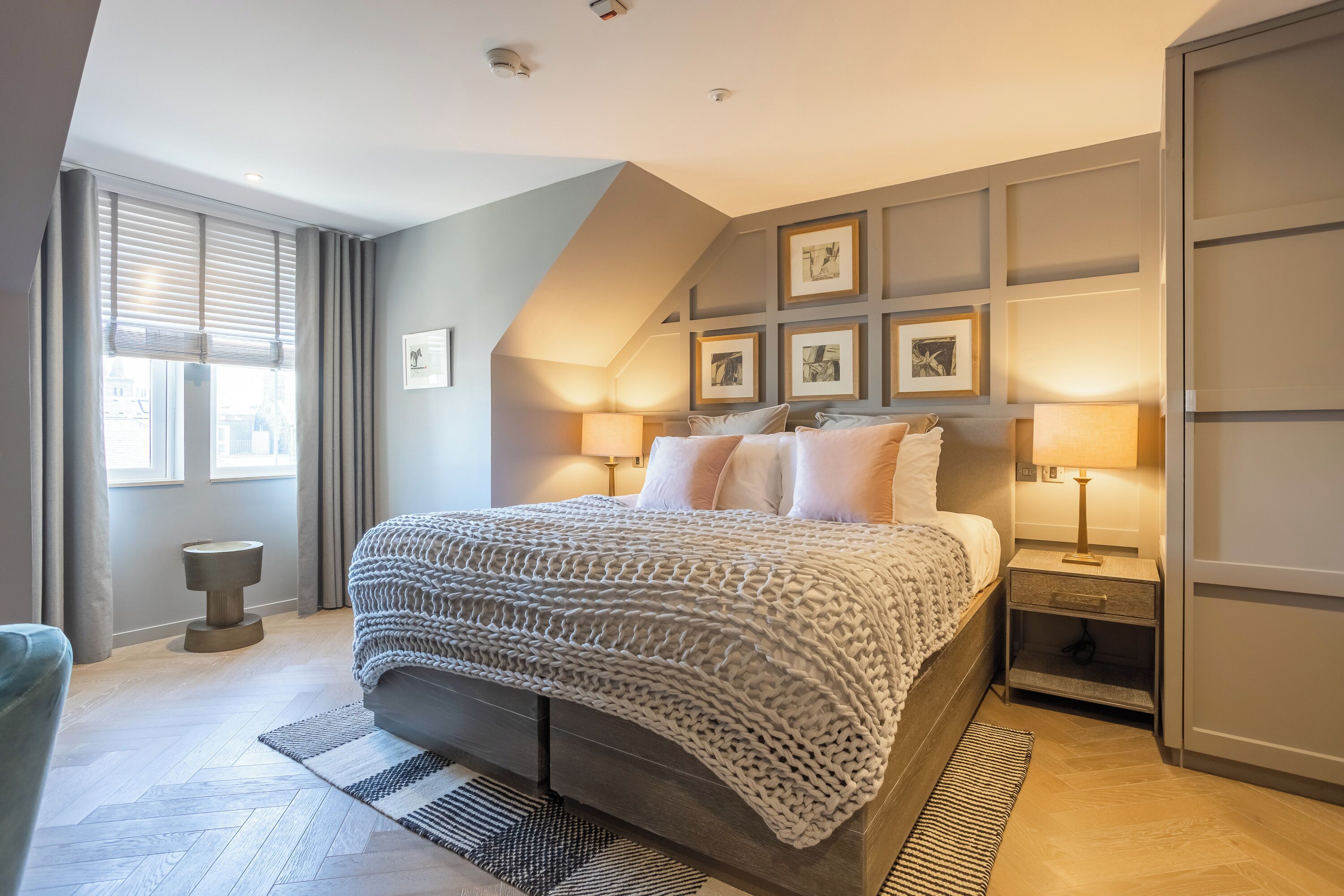 Un dormitorio moderno con una cama grande, dos mesitas de noche con lámparas, una pared gris con cuadros enmarcados y una ventana con cortinas.