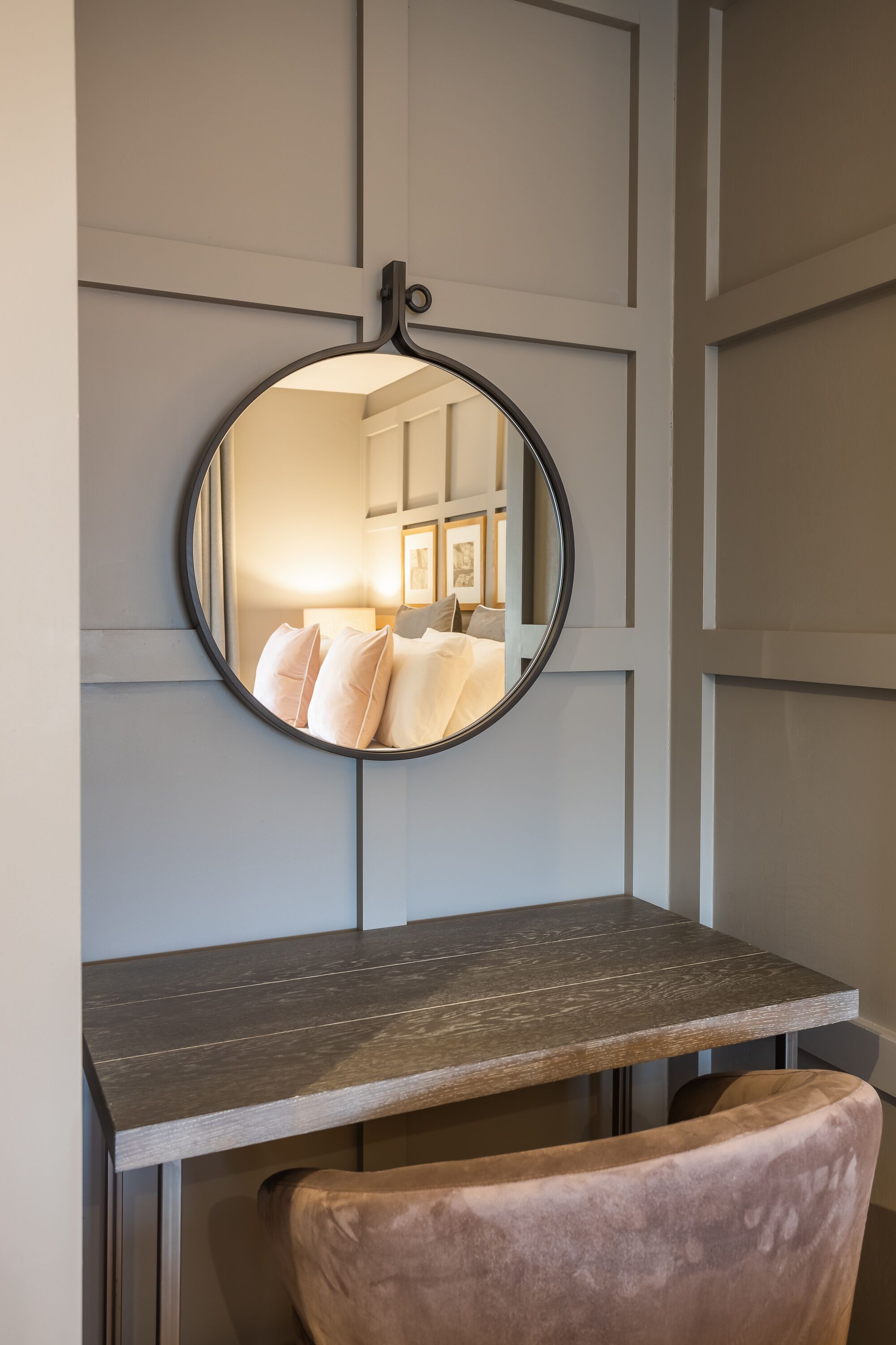 A round mirror on a wall, reflecting a room with a bed, pillows, and a lamp.