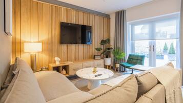 A modern living room with a sofa, a round coffee table, a TV mounted on a wooden wall, and a large window with a view.