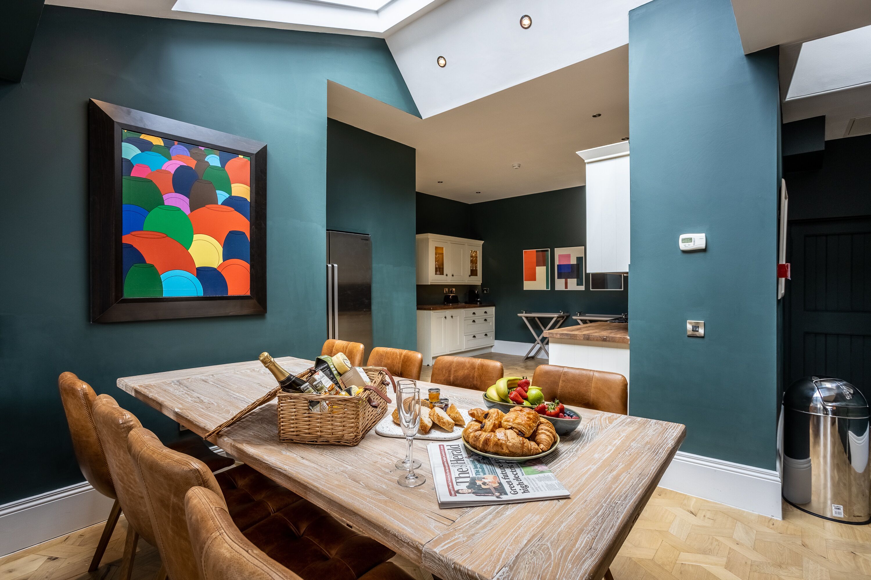 Un coin repas avec une table en bois dressée pour le repas, un tableau abstrait aux couleurs vives accroché au mur et une vue sur la cuisine.