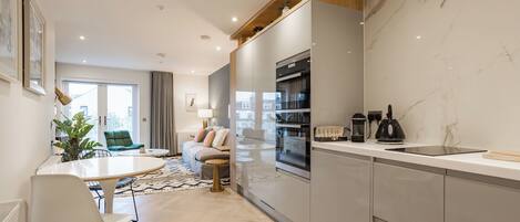 A modern kitchen with a built-in oven, a sink, and a countertop. There is a dining area with a table and chairs, and a sofa in the background.