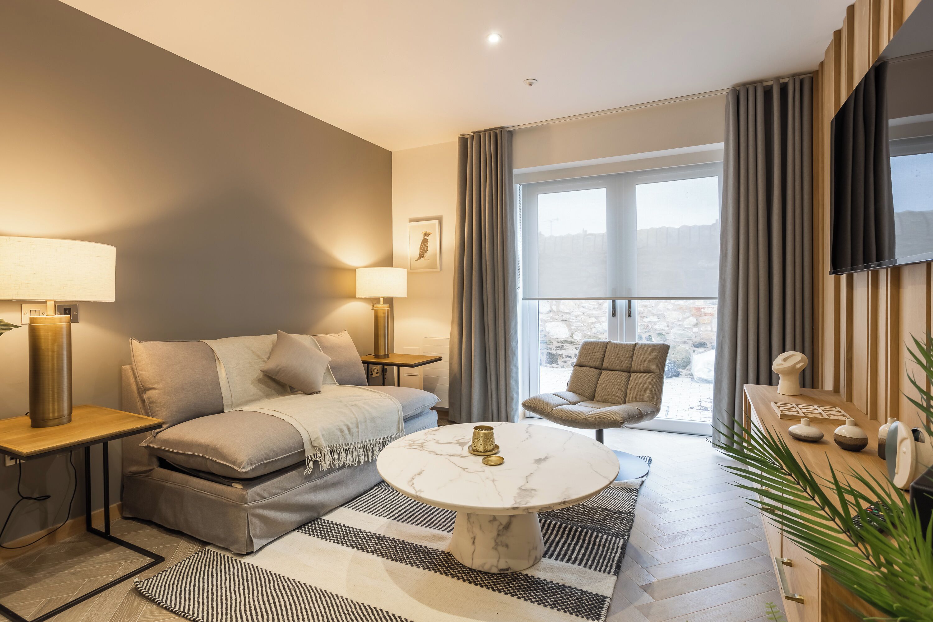 A modern living room with a sofa, a round coffee table, a TV mounted on the wall, and a large window with curtains.