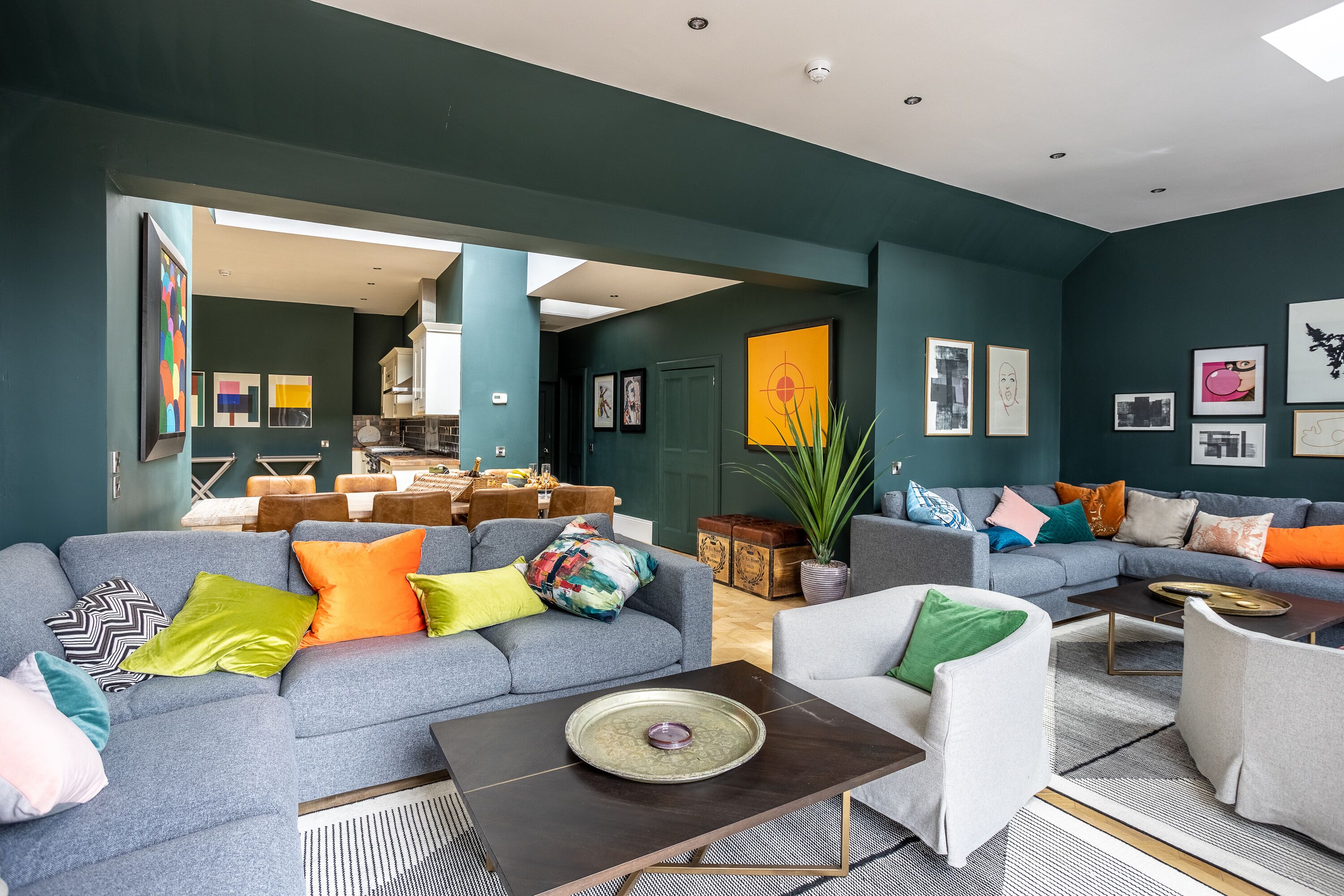 Una sala de estar moderna con un sofá seccional gris, un sillón blanco y una mesa de centro. Hay cojines de colores y una planta en maceta.