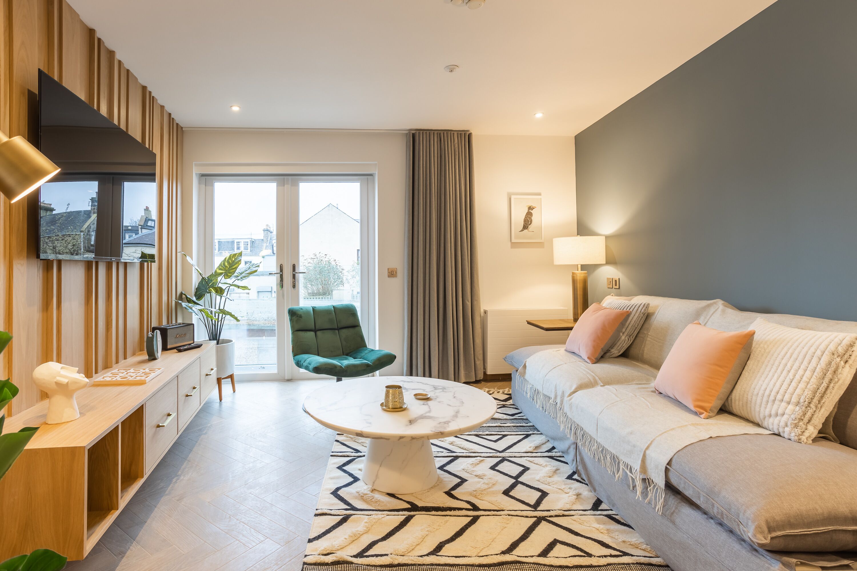 Ein modernes Wohnzimmer mit einer Couch, einem Couchtisch, einem an der Wand befestigten Fernseher und Blick ins Freie durch eine Glastür.