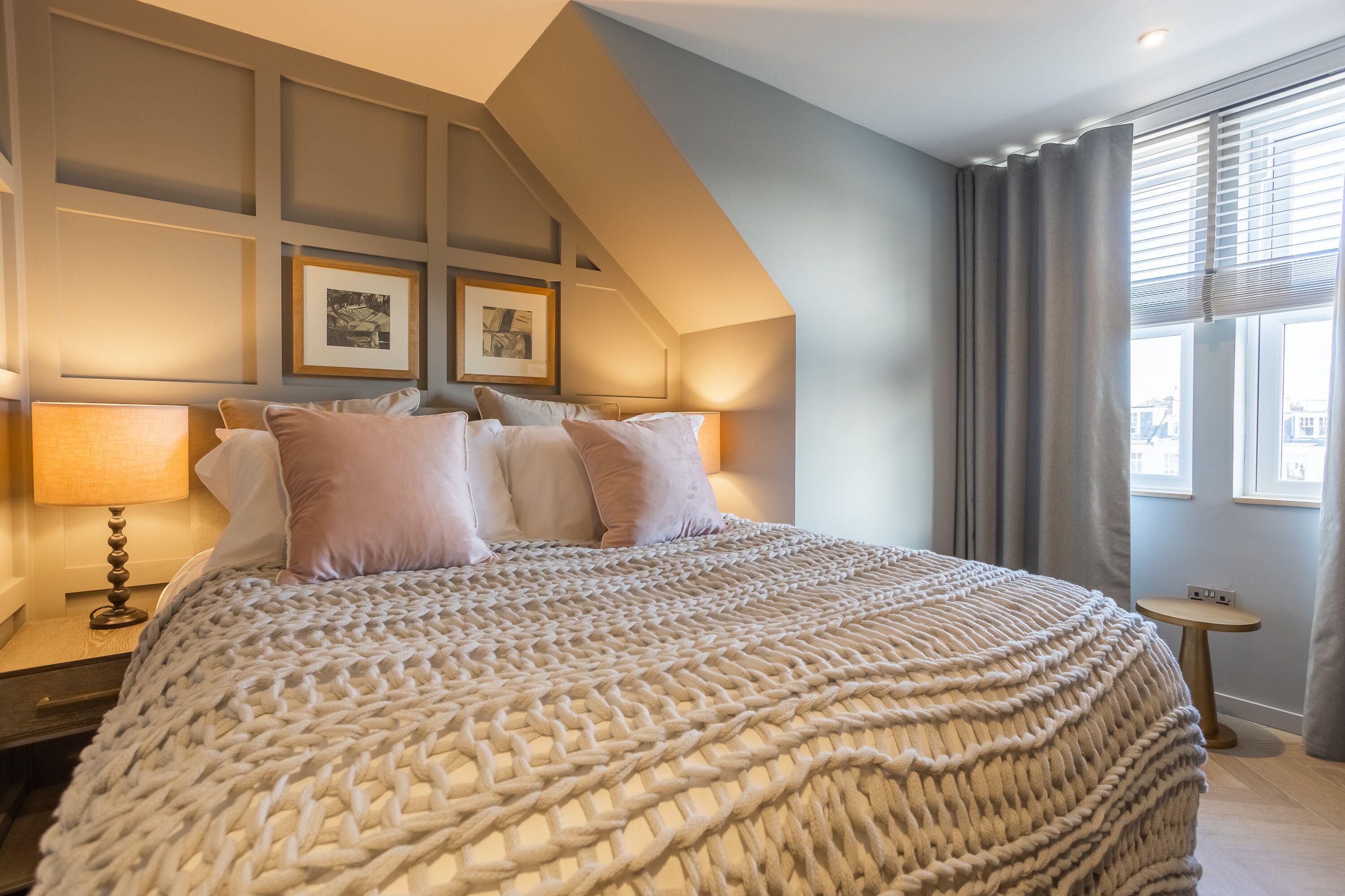 Un dormitorio con una cama grande, una mesita de noche con lámpara, un cuadro en la pared y una ventana con cortinas.