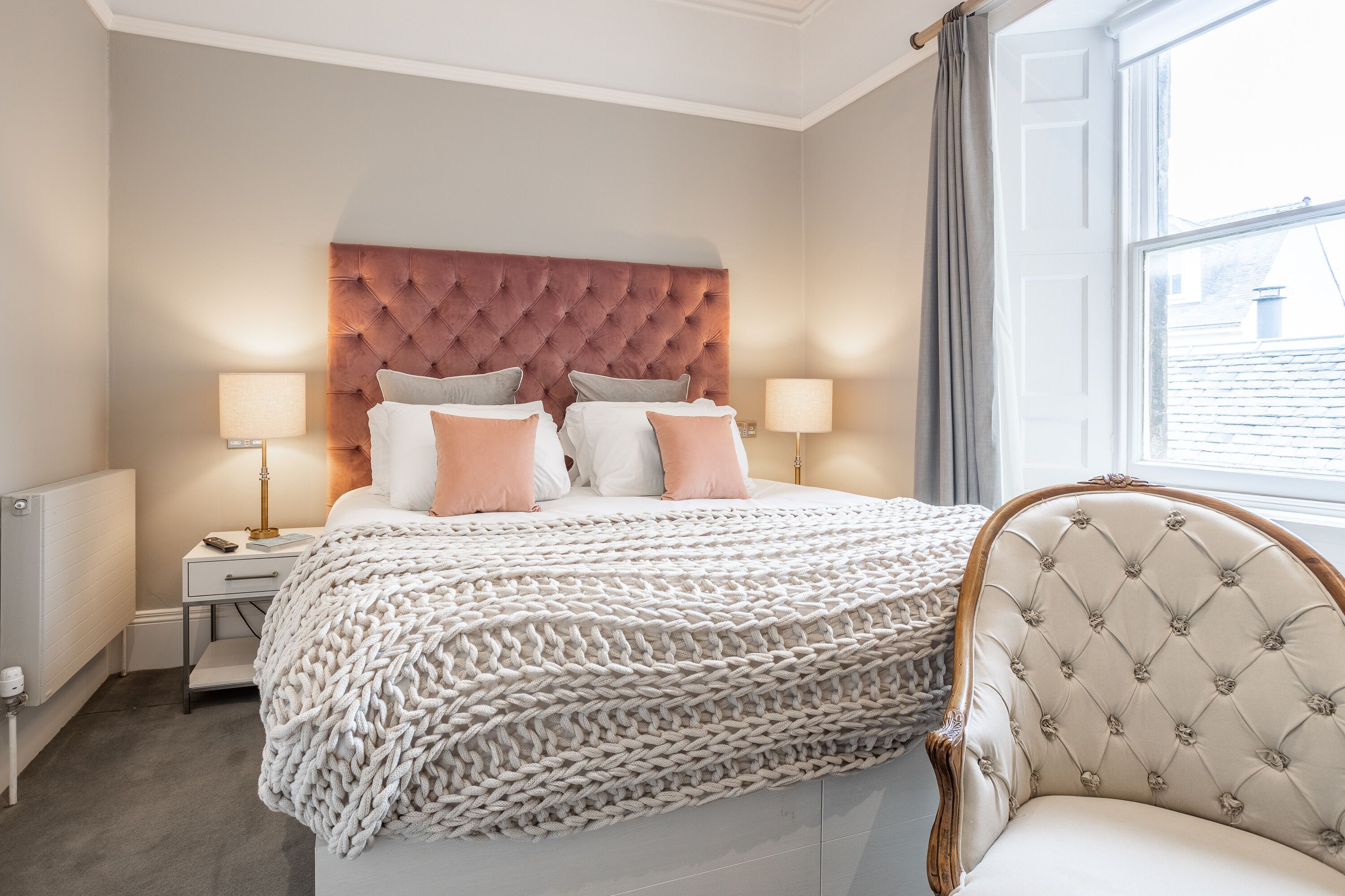 A bedroom with a tufted headboard, two bedside tables with lamps, a large bed with a chunky knit blanket, and a chair with a floral pattern.