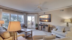 A spacious living room with a ceiling fan, a television mounted on the wall, a bookshelf, and a view of the outdoors through large windows. - 3305 Windsor Court South -Amazing renovated 3rd Floor Oceanfront Villa (Hilton Head Island)