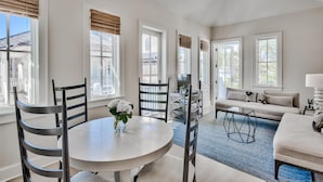 Ein geräumiges Wohnzimmer mit rundem Tisch, zwei Stühlen, einer Couch und einem Fernseher. Große Fenster mit Jalousien lassen viel Tageslicht herein.