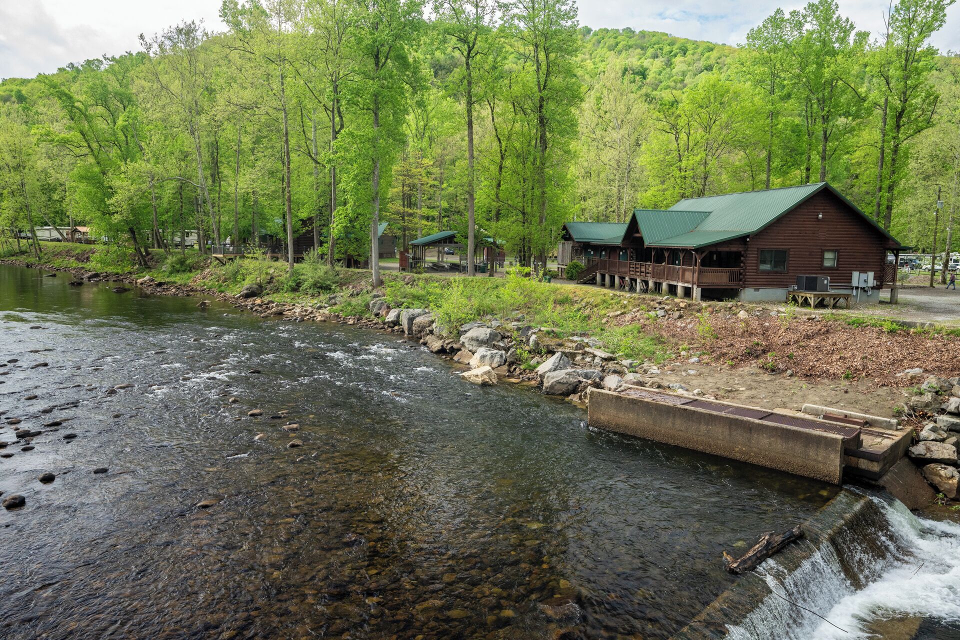 A lodge by a river with a wooden structure, a deck, and a view of the surrounding forest.
