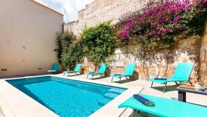 Une piscine à l’eau d’un bleu cristallin, entourée de chaises longues et bordée d’un mur de pierre orné de plantes fleuries.